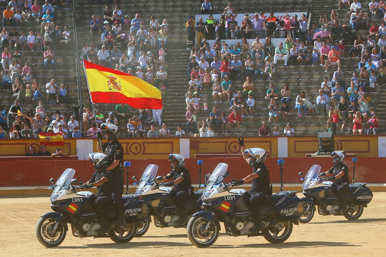 Se muestran cinco policías motorizados.  La primera moto la ocupan dos policías, portando uno de ellos la bandera española.