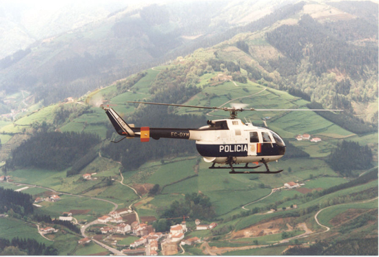 Helicóptero de Policía Nacional en vuelo realizando una patrulla aérea sobre una población ubicada en una zona de montes y prados.