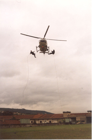 Helicóptero de Policía Nacional en vuelo de poca altura con dos miembros de Unidades Especiales en el exterior, iniciando descenso en rapel.