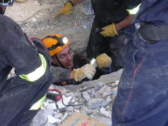Miembro de un equipo de rescate se introduce en agujero, ayudado por otras personas, alrededor se pueden observar multitud de escombros.
