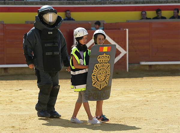 Dos niñas portando escudo y casco de la policía. Detrás policía con traje para desactivación de explosivos. Andan sobre el ruedo de la plaza de toros.