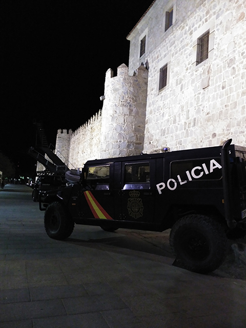 Vehículo policial rotulado de la UIP, URO Vamtac S3, frente a la muralla de Ávila. Es de noche, la muralla se encuentra iluminada.