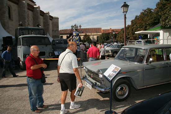 Dos visitantes de la exposición abierta observando un vehículo antiguo de la Policía, modelo Seat 1500 de color gris.