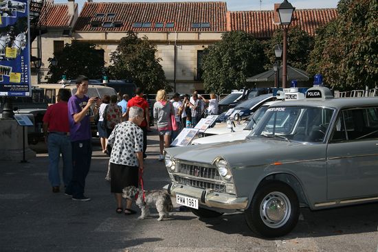 Varios visitantes de la exposición abierta observando un vehículo antiguo de la Policía, modelo Seat 1500 de color gris y otros vehículos más.