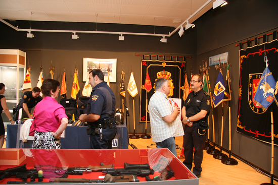 Varios visitantes y Policías Nacionales en un sala de la exposición donde hay banderines de varias unidades y vitrinas con armas.