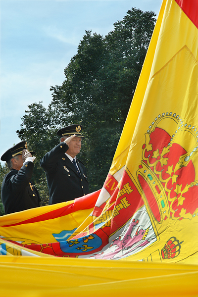Una bandera de España de grandes dimensiones y dos Policías Nacionales de uniforme saludando a la bandera.