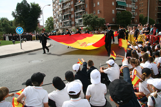 Varios Policías Nacionales portan extendida una bandera de España de grandes dimensiones mientras niños y ciudadanos asisten al acto.