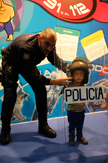 Niño pequeño posando con un casco y escudo policial, sujeto por un agente de policía.