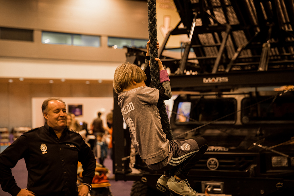 Niño intentando trepar por una cuerda junto a un agente que vigila por su seguridad, al fondo vehículo del Grupo Especial de Operaciones