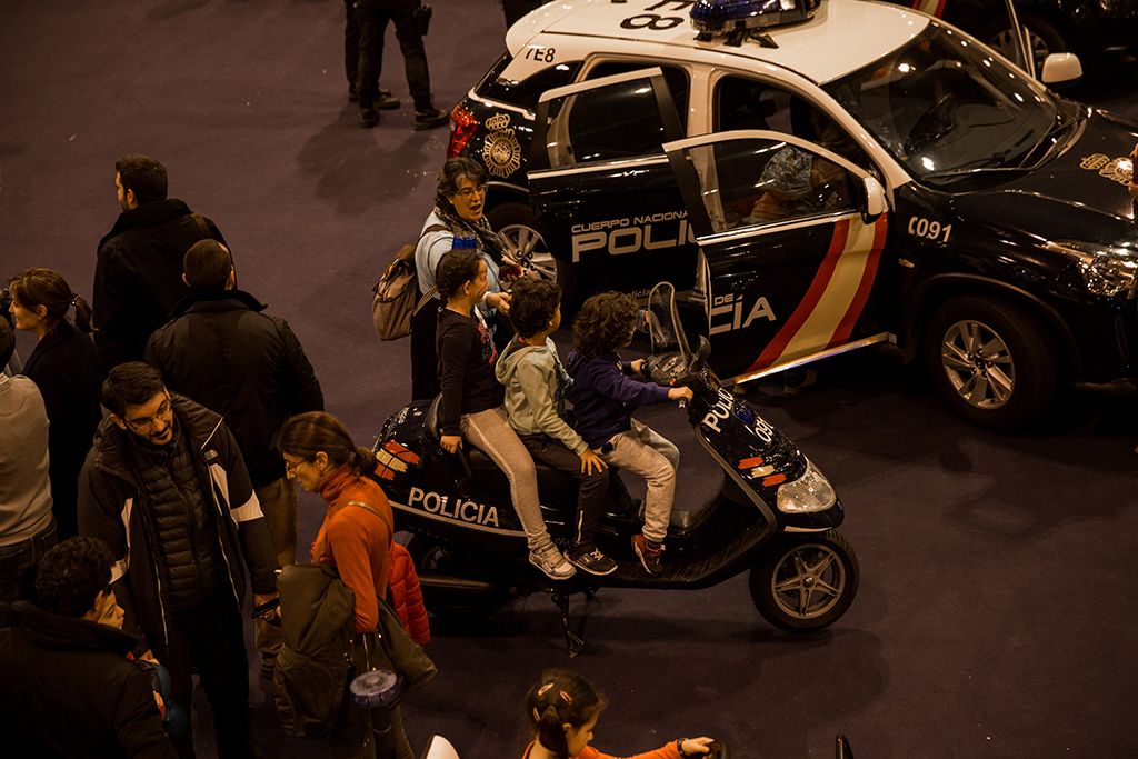 Tres niños subidos en una motocicleta, junto a un coche policial y varios adultos.