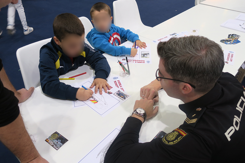 Dos niños coloreando dibujos en una mesa y un agente de policía ayudándoles.