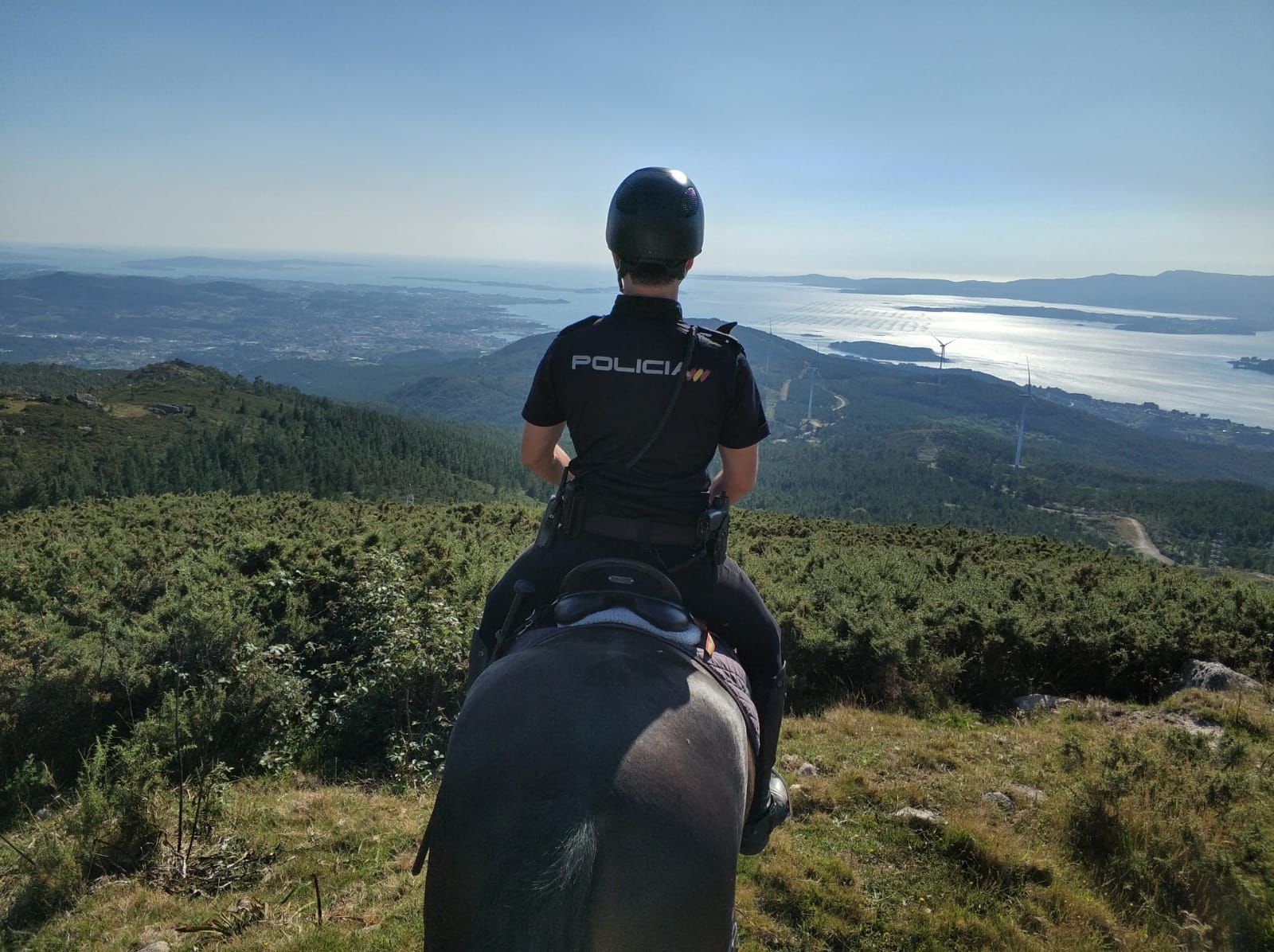 Un policía montado a caballo de espaldas, en lo alto de un monte desde el que se observa el mar y las montañas
