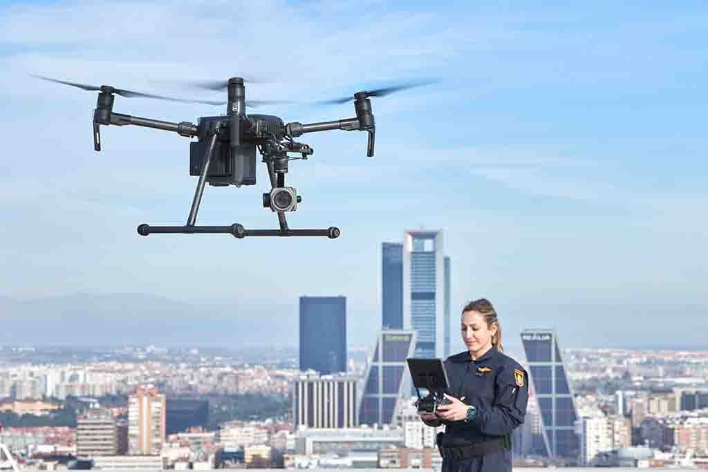 Mujer uniformada del Servicio de Medios Aéreos pilotando un dron con cámara. De fondo varios rascacielos de la ciudad de Madrid.