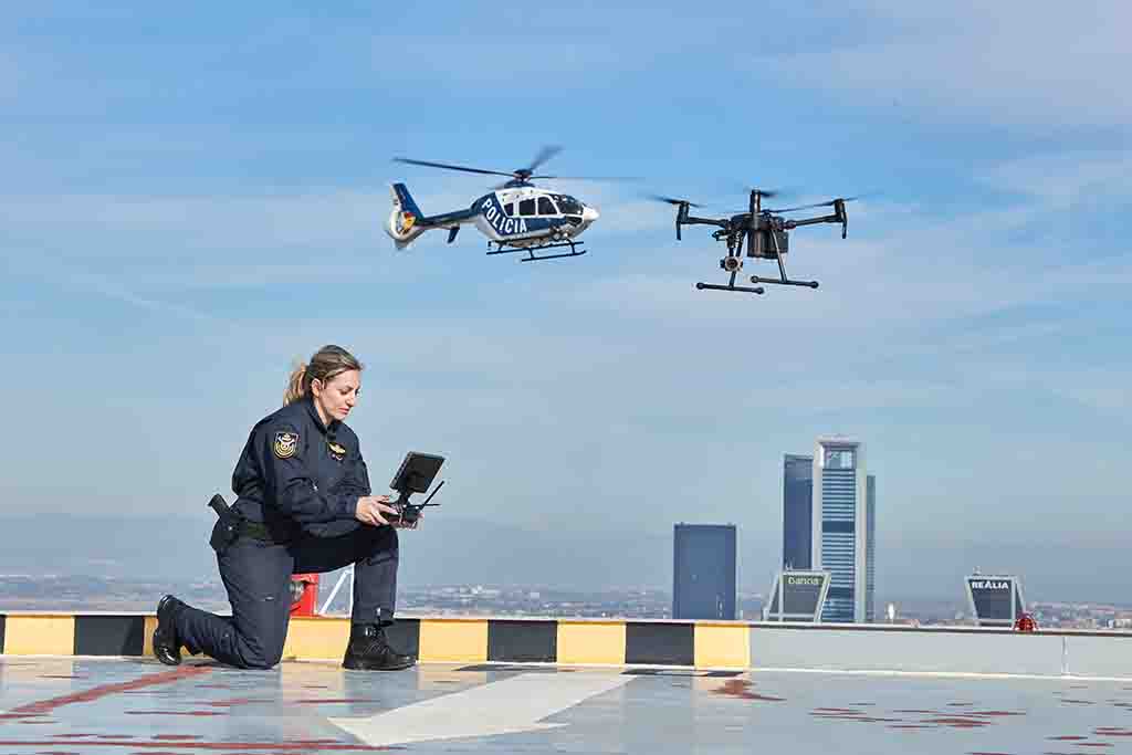 Mujer uniformada del Servicio de Medios Aéreos pilotando un dron. De fondo, rascacielos de la ciudad de Madrid y el helicóptero de la Policía Nacional