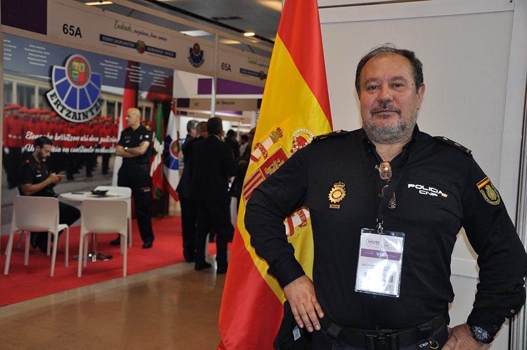 Inspector Jefe de la Policía Nacional posando con la bandera de España. De fondo se ve el stand de la Ertzaintza.